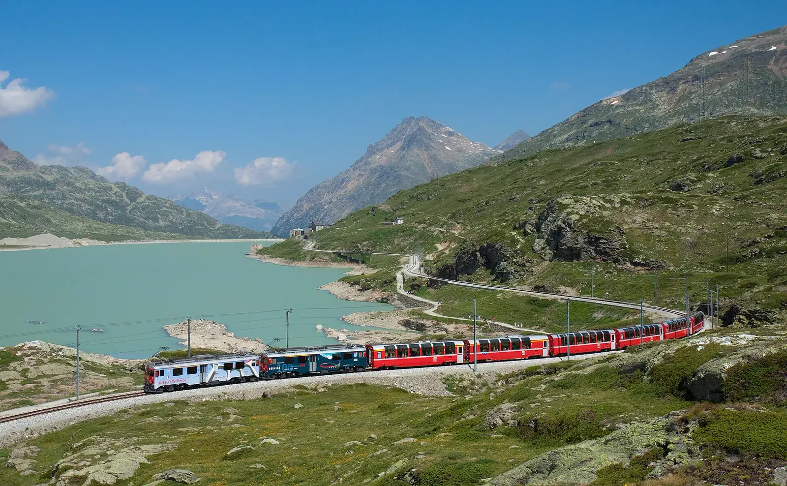 A red Bernina Express train travelling beside the turquoise Lago Bianco under a bright blue alpine sky